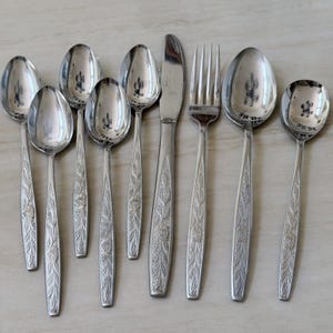 Puede incluir: Un juego de nueve piezas de cubiertos de acero inoxidable que incluye cucharas, un cuchillo y un tenedor. Cada mango presenta un patrón floral en relieve con hojas y una rosa. Los utensilios tienen un acabado plateado pulido.