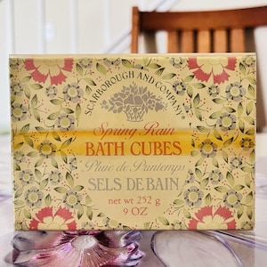 May include: A box of Scarborough and Company Spring Rain bath cubes. The box features a floral pattern with pink, green, and grey flowers on a cream background. The text "BATH CUBES" is prominent, and the net weight is 252 g.