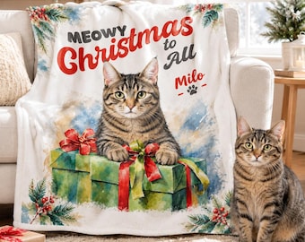 May include: A white fleece blanket featuring a watercolor-style illustration of a tabby cat resting on a green gift box with a red bow. The text reads Meowy Christmas to All and Milo, accompanied by a paw print, holly, and pine branch accents.