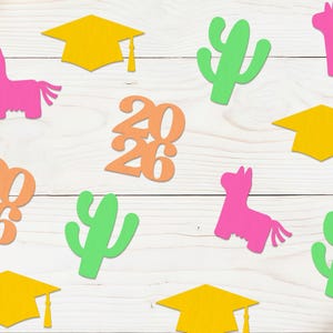 May include: A collection of colorful cutouts on a white wooden surface. The cutouts include pink pinatas, yellow graduation caps, green cacti, and the numbers 2026 in orange. The arrangement suggests a graduation or celebratory theme.