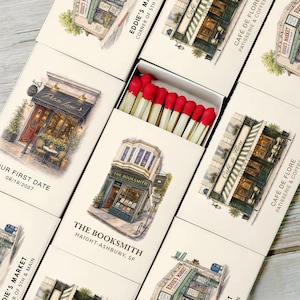 May include: A collection of rectangular matchboxes featuring watercolor illustrations of storefronts including The Booksmith, Cafe de Flore, and Eddie's Market. One open matchbox reveals wooden matches with red tips. The boxes display text such as Our First Date 09/19/2027.