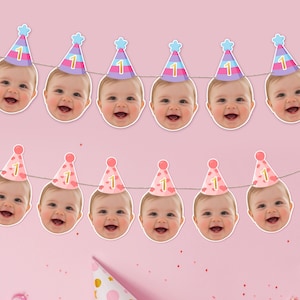 May include: A festive banner featuring smiling baby faces wearing party hats with the number "1". The hats are in pastel colors of pink, blue, and purple. The banner is strung on a thin white string against a pink background.