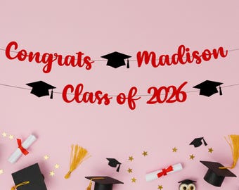 May include: A pink graduation banner with the words "Congrats Madison Class of 2026" in red script. Black graduation caps are strung along the banner. The background is pink with scattered graduation-themed decorations.