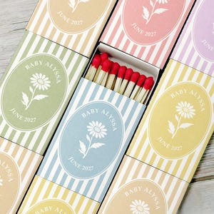 May include: A collection of rectangular matchboxes featuring vertical stripes in various pastel colors. Each box displays an oval emblem with a white daisy illustration, the text BABY ALYSSA, and the date JUNE 2027. One box is open, revealing red-tipped matches.
