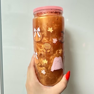 May include: A cylindrical glass tumbler filled with a swirling metallic copper liquid, topped with a pink lid and orange glitter. The exterior features small decals of orange pumpkins with white bows, yellow maple leaves, a pink sweater, and boots.
