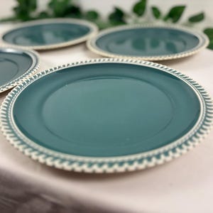 Peut inclure: Quatre assiettes en céramique de couleur teal avec une bordure blanche texturée. Les assiettes sont disposées sur une surface de couleur claire, avec du feuillage vert en arrière-plan. Les assiettes sont de couleur teal unie.
