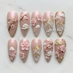 May include: A set of ten almond-shaped artificial nails. Each nail features a light pink base with 3D floral and mountain designs in white, gold, and pink. The designs include cherry blossoms, clouds, and mountain ranges.