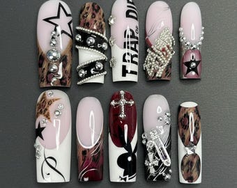 Op de afbeelding: Een set van tien kunstnagels met diverse ontwerpen. De nagels hebben een mix van luipaardprint, sterren, kruisen en Playboy bunny-motieven, samen met strass-steentjes en de tekst "TRAP DIE". De nagels zijn in tinten wit, roze, zwart en bordeaux.