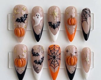 Może przedstawiać: Zestaw dziesięciu migdałowych sztucznych paznokci z motywami halloweenowymi. Paznokcie ozdobione są wypukłymi pomarańczowymi dyniami 3D, białymi duchami, czarnymi nietoperzami i misternymi czarnymi pajęczynami na cielistym tle ze złotymi gwiazdkami.