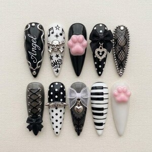 May include: A set of ten press-on nails featuring diverse designs including black and white polka dots, a skull with musical notes, a pink paw print, a black bow with a heart charm, lace patterns, corset lacing, and piano keys.