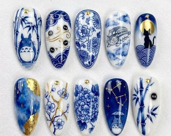 May include: A set of ten almond-shaped press-on nails featuring blue and white porcelain-inspired designs. The patterns include bamboo, floral motifs, soot sprites, a cat silhouette, a bus, and gold leaf accents, each topped with a small gold stud.