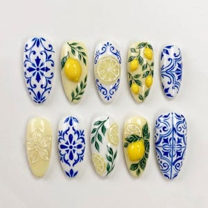 Può includere: Un set di dieci unghie finte a forma di mandorla con vari design, tra cui motivi a piastrelle blu e bianche, limoni gialli in rilievo con foglie verdi, dettagli di fette di limone e un singolo fiore in rilievo su una base giallo pallido.