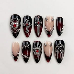 May include: A set of ten press-on nails featuring a gradient of black to deep red. The designs include silver metal charms such as a heart with barbed wire, a cross, a sword, horns, spikes, an eye with circuit lines, and a gothic archway.