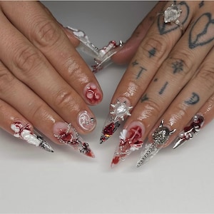 May include: Close-up of a set of long, pointed acrylic nails. The nails feature a variety of designs, including red and white accents, silver embellishments, and a clear base. Some nails have a blood-like effect.