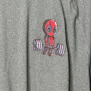 May include: A heather gray long-sleeve shirt featuring an embroidered graphic of a cartoon character with a red mask and suit lifting a heavy barbell with large gray weights on each side. The fabric has a textured, speckled appearance throughout.