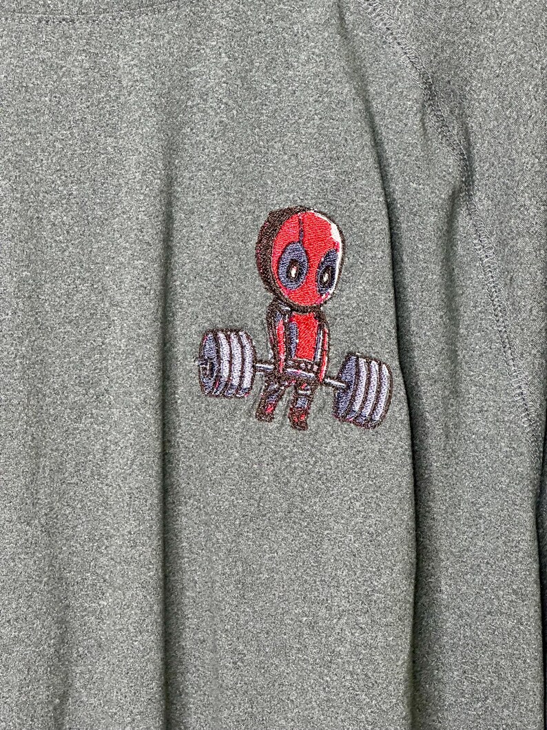 May include: A heather gray long-sleeve shirt featuring an embroidered graphic of a cartoon character with a red mask and suit lifting a heavy barbell with large gray weights on each side. The fabric has a textured, speckled appearance throughout.