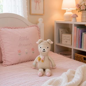 May include: A beige knit stuffed animal with a small pink scarf and pink accents on the ears and feet. The toy has embroidered facial features and sits on a pink quilted bedspread featuring a pillow with the text little dreamer and flowers.