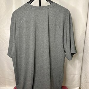 May include: A short-sleeved, heather gray t-shirt with a crew neckline and raglan sleeves. The garment is displayed on a black hanger, showing the back view of the shirt with a solid, textured fabric finish and a simple, clean construction.