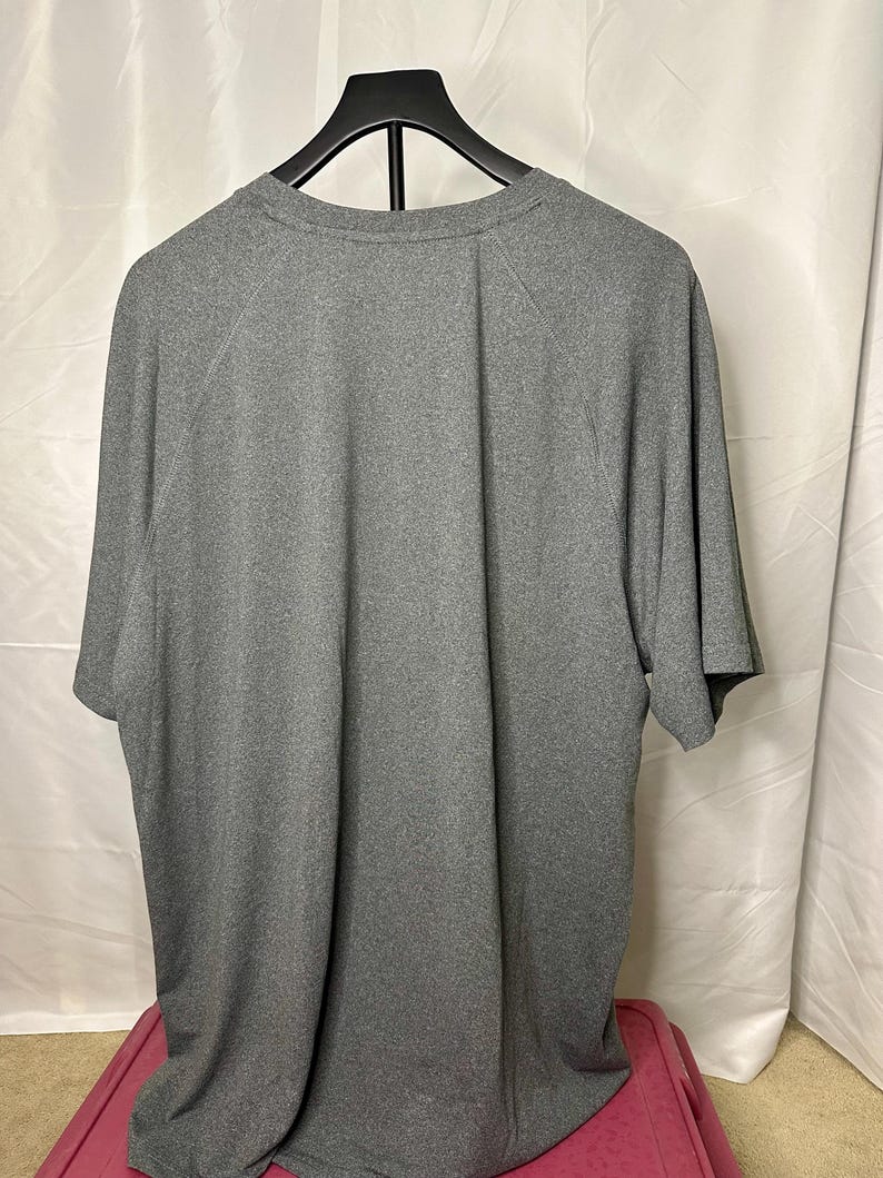 May include: A short-sleeved, heather gray t-shirt with a crew neckline and raglan sleeves. The garment is displayed on a black hanger, showing the back view of the shirt with a solid, textured fabric finish and a simple, clean construction.