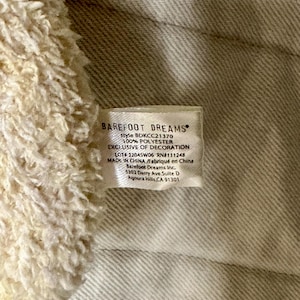 May include: Close-up of a tag from Barefoot Dreams. The tag is white with black text, including the brand name, style number, and material composition. The tag also includes the text "EXCLUSIVE OF DECORATION" and the address of the company.