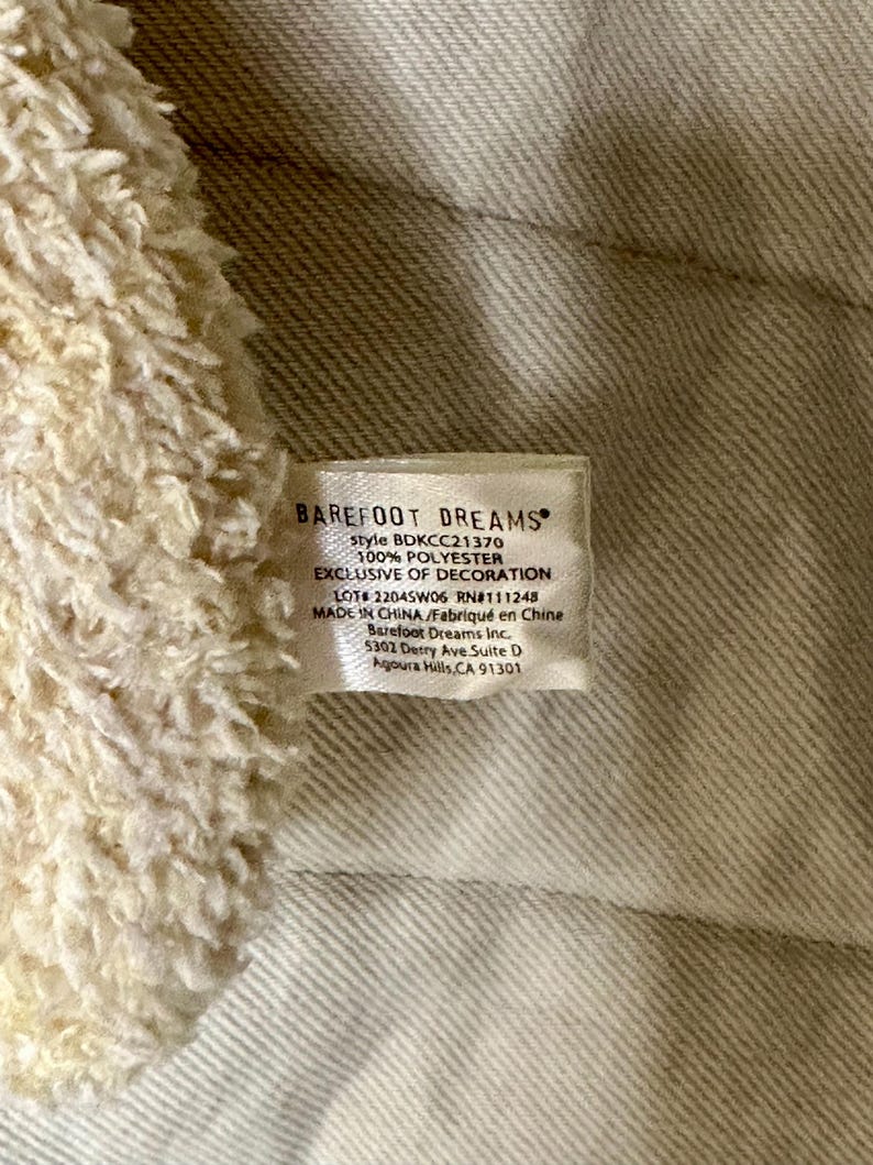 May include: Close-up of a tag from Barefoot Dreams. The tag is white with black text, including the brand name, style number, and material composition. The tag also includes the text "EXCLUSIVE OF DECORATION" and the address of the company.