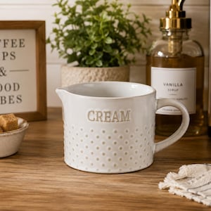 May include: A white ceramic creamer pitcher featuring a textured polka dot pattern on the lower half and the word CREAM embossed in capital letters on the front. The pitcher has a small pouring spout and a curved handle on the right side.