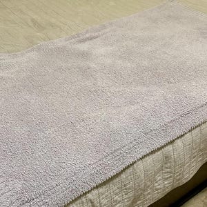 Pu&ograve; includere: Una coperta rettangolare color lavanda chiaro con una superficie testurizzata e granulosa, dotata di un bordo a coste lungo i lati. La coperta &egrave; piegata e distesa, mostrando un colore viola tenue e una densa lavorazione a maglia.