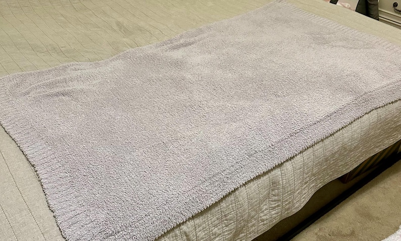 Pu&ograve; includere: Una coperta rettangolare color lavanda chiaro con una superficie testurizzata e granulosa, dotata di un bordo a coste lungo i lati. La coperta &egrave; piegata e distesa, mostrando un colore viola tenue e una densa lavorazione a maglia.
