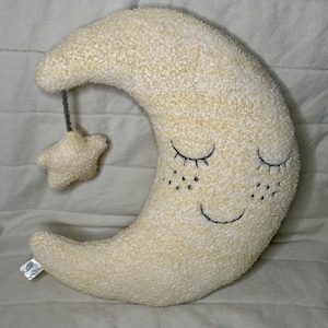 May include: A plush, cream-colored crescent moon pillow with embroidered closed eyes and a smiling mouth. A small star is suspended from the moon by a gray string. The pillow has a soft, textured surface.