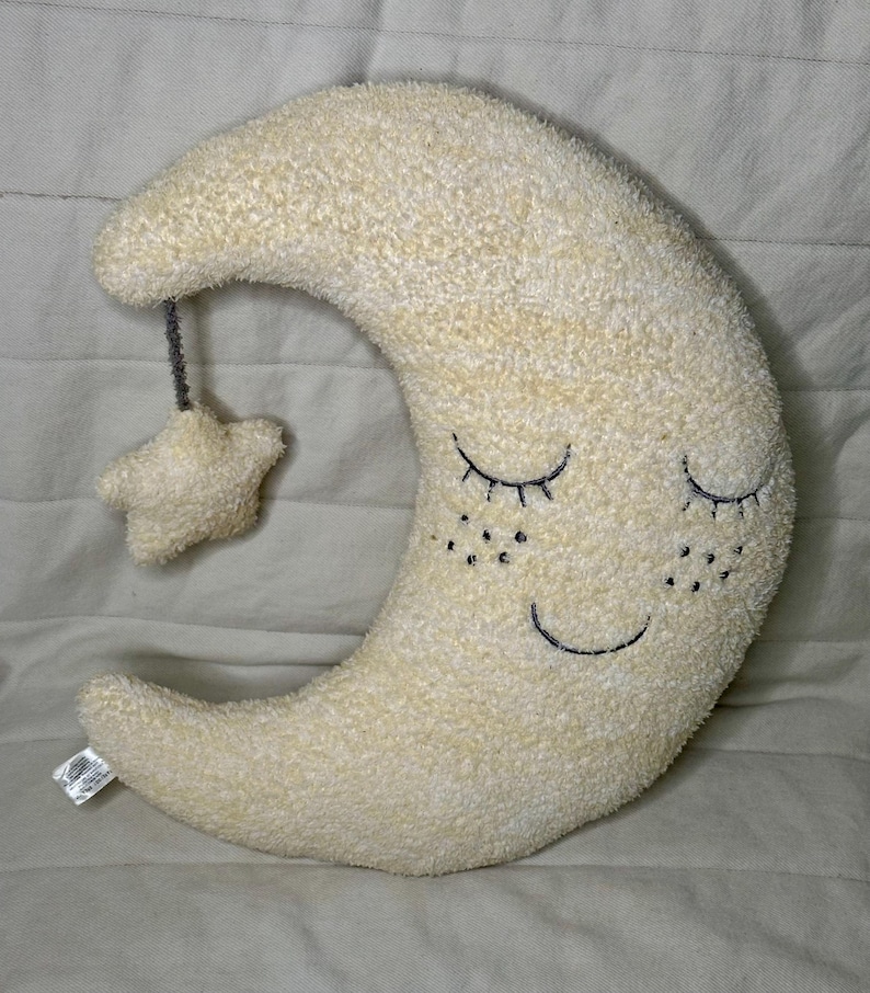 May include: A plush, cream-colored crescent moon pillow with embroidered closed eyes and a smiling mouth. A small star is suspended from the moon by a gray string. The pillow has a soft, textured surface.
