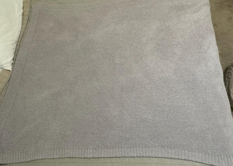 Pu&ograve; includere: Una coperta lavorata a maglia color grigio lavanda chiaro con una consistenza morbida e soffice. La coperta presenta un ampio bordo a coste lungo i lati e un motivo a maglia denso e uniforme su tutto il corpo principale, creando un colore.