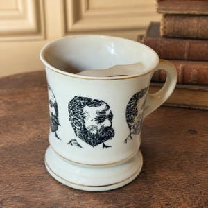 May include: A cream-colored ceramic shaving mug featuring a black ink illustration of a bearded man in profile. The mug has a wide handle, a flared base, and a gold-toned rim. An interior ledge is visible inside the top of the mug.