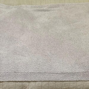 Pu&ograve; includere: Una coperta rettangolare color lavanda chiaro con una superficie testurizzata e soffice, dotata di un bordo in maglia a coste lungo i lati. La coperta presenta una piccola etichetta rettangolare in tessuto bianco nell'angolo in basso a destra.