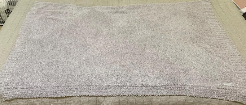 Pu&ograve; includere: Una coperta rettangolare color lavanda chiaro con una superficie testurizzata e soffice, dotata di un bordo in maglia a coste lungo i lati. La coperta presenta una piccola etichetta rettangolare in tessuto bianco nell'angolo in basso a destra.