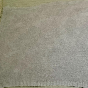 Pu&ograve; includere: Una coperta color lavanda chiaro a tinta unita con una consistenza morbida, soffice e felpata. La coperta presenta un bordo a coste distinto lungo i lati, che fornisce una cornice strutturata al design tessile monocromatico e quadrato.