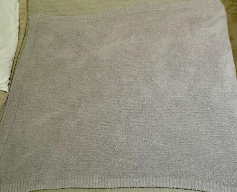 Pu&ograve; includere: Una coperta color lavanda chiaro a tinta unita con una consistenza morbida, soffice e felpata. La coperta presenta un bordo a coste distinto lungo i lati, che fornisce una cornice strutturata al design tessile monocromatico e quadrato.