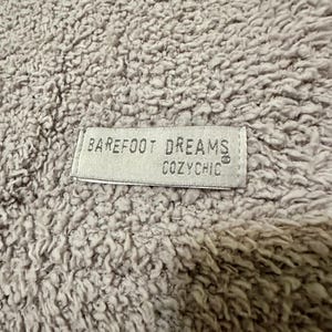 Pu&ograve; includere: Un tessuto testurizzato a pelo lungo di colore beige chiaro con un'etichetta rettangolare in tessuto cucita sulla superficie. L'etichetta &egrave; beige chiaro con testo scuro che recita BAREFOOT DREAMS COZYCHIC, con un piccolo simbolo di marchio registrato.