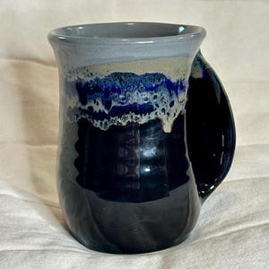 Puede incluir: Una taza de cerámica hecha a mano con un asa curva única. La taza presenta un degradado de colores, comenzando con un azul marino oscuro en la base, pasando a azules más claros y blanco cerca del borde. El borde es gris claro. La taza tiene un acabado brillante.