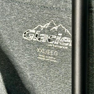 May include: A heathered dark gray t-shirt featuring a white printed logo of a mountain range above the text Glacier performance. The interior neck label displays the size XXL/EEG and the text Made in China and Hecho en China.