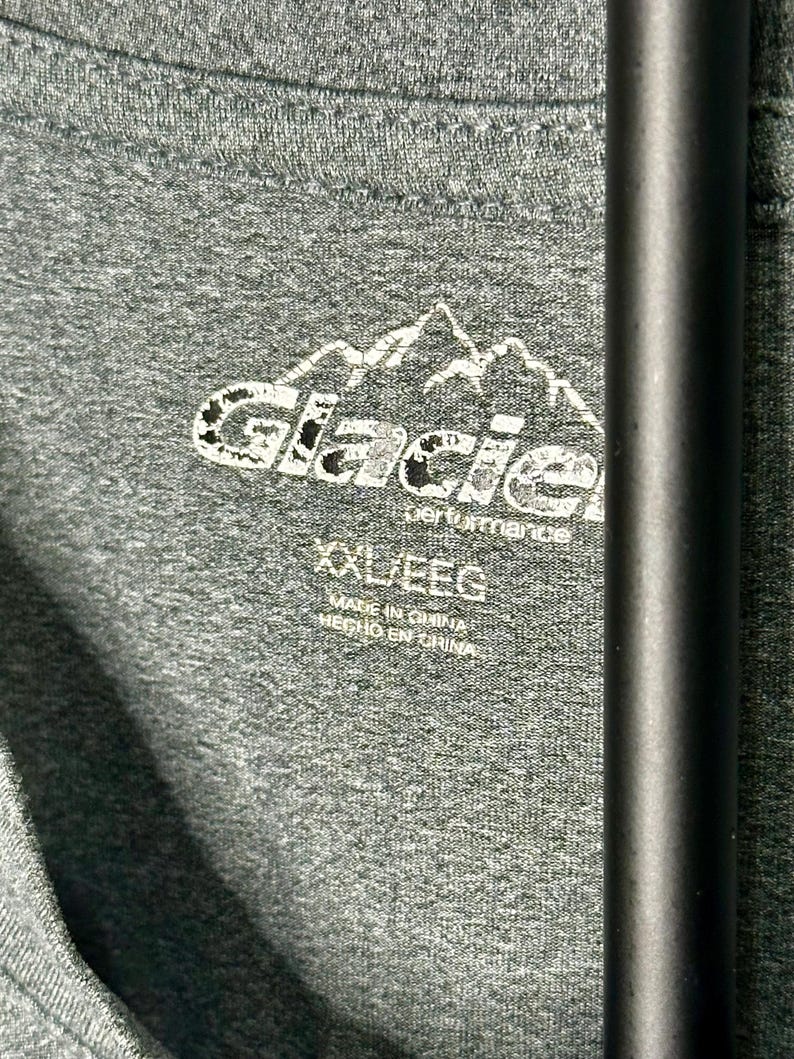 May include: A heathered dark gray t-shirt featuring a white printed logo of a mountain range above the text Glacier performance. The interior neck label displays the size XXL/EEG and the text Made in China and Hecho en China.