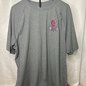 May include: A heather gray short-sleeve t-shirt featuring a small graphic on the upper left chest. The graphic depicts a red-masked character lifting a barbell with gray weights. The shirt has a crew neckline and a standard athletic fit.
