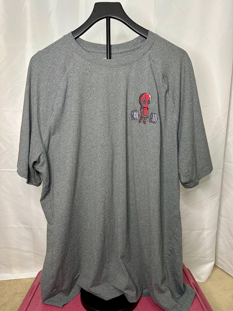 May include: A heather gray short-sleeve t-shirt featuring a small graphic on the upper left chest. The graphic depicts a red-masked character lifting a barbell with gray weights. The shirt has a crew neckline and a standard athletic fit.