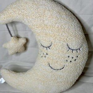 May include: A plush, cream-colored crescent moon pillow with embroidered closed eyes, freckles, and a smiling mouth. A small star is attached by a gray string. The pillow is soft and fluffy.
