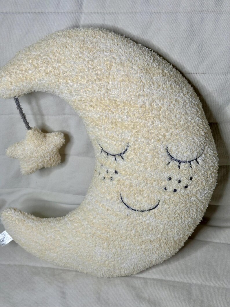 May include: A plush, cream-colored crescent moon pillow with embroidered closed eyes, freckles, and a smiling mouth. A small star is attached by a gray string. The pillow is soft and fluffy.