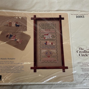 Op de afbeelding: Een 1693 Family Sampler kruissteekpakket van The Creative Circle in plastic verpakking. De set bevat taupekleurige Aida-stof, borduurgaren, een naald en instructies voor het maken van een sampler met een alfabet, huis en familiefiguren.