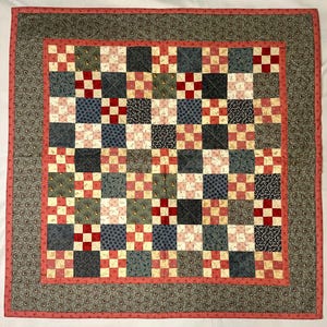 Puede incluir: Una colcha de patchwork cuadrada con una cuadrícula de bloques alternos en tonos azul marino, crema y rojo. El patrón central está enmarcado por un borde de color coral sólido y un borde exterior de tela gris oscuro con estampado floral.