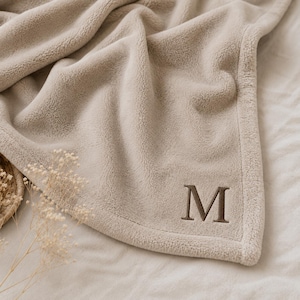 May include: A soft, beige fleece blanket featuring a plush, textured surface and a finished hem. The blanket is embroidered with a single, dark brown capital letter M in a serif font positioned near the bottom right corner of the fabric.