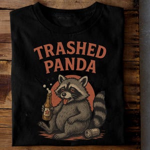 May include: Black t-shirt with a cartoon raccoon design. The raccoon is holding a beer bottle and a can, with the text "Trashed Panda" above in orange. The raccoon has a red tongue sticking out.