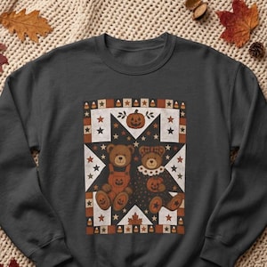 May include: A dark gray crewneck sweatshirt featuring a central graphic of two teddy bears surrounded by a star and pumpkin pattern. The design includes a jack-o'-lantern, candy corn, and maple leaves in shades of orange, brown, and cream.