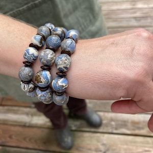 May include: A stack of three beaded bracelets. The bracelets feature large, round beads in shades of blue, white, and brown, with a marbled effect. Dark brown spacers separate the beads. The bracelets are worn on a wrist.